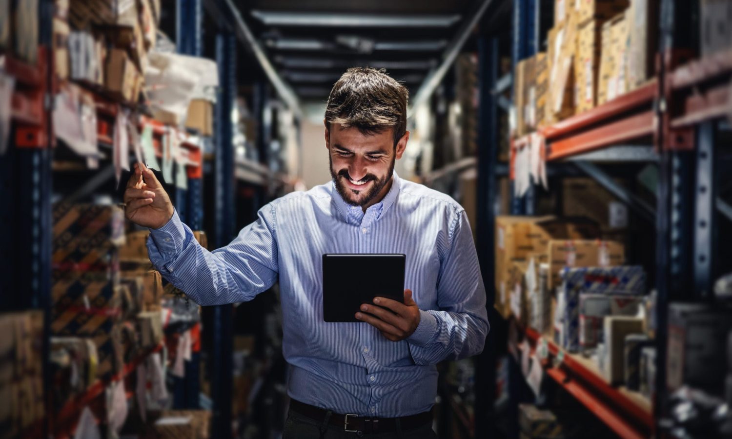 Smiling bearded supervisor standing in warehouse full of boxes and checking on goods ready for deliver.